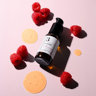 Bottle of Raspberry Seed Serum with raspberries and drops of the beauty oil on a pink background