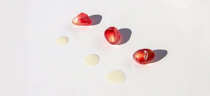 Close up shot of Pomegranate Eye Serum bottle and 3 drops of the product on a beige backround 