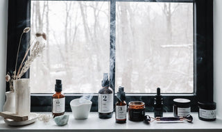 Skincare products sitting on a windowsill with inscence and snow outside the window