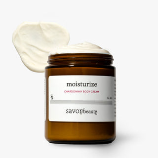 An amber glass jar of Savor Beauty moisturize Chardonnay Body Cream with a white label, shown with a dollop of the white cream on top against a white background.