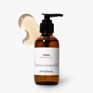 An amber glass pump bottle labeled 'relax CINNAMON ARNICA OIL' by Savor Beauty, shown on a clean white background with a beige cream smear behind it.