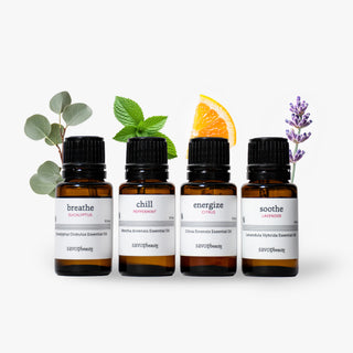 Four amber glass Savor Beauty essential oils bottles with black caps and white labels reading breathe, chill, energize, and soothe, arranged on a white background with eucalyptus leaves, mint, an orange slice, and lavender sprigs.