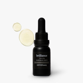 A black dropper bottle of SAVOr beauty brilliance Fountain of Youth serum, shown on a light gray background with two circular droplets of the pale yellow serum floating to the left.