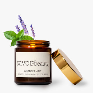 An amber glass jar candle with a white label reading 'savor beauty' and 'LAVENDER MINT,' shown with its metallic gold lid leaning against it on a light gray background.