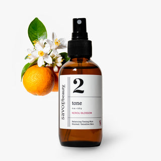 An amber glass spray bottle labeled 'savor beauty tone neroli blossom' stands on a light gray background, styled with an orange, green leaves, and white neroli blossoms on the left side.
