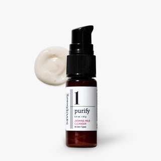 A small amber glass pump bottle labeled '1 purify' by Savor Beauty, shown on a light gray background with a dollop of creamy off-white cleanser beside it.