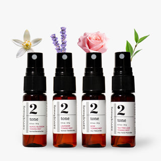 Four amber spray bottles labeled '2 tone' by Savor Beauty are arranged in a row on a white background, each topped with a different botanical sprig: neroli blossom, lavender, pink rose, and tea tree leaves.