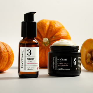 Savor Beauty Pumpkin Hydra Glow Duo skincare products displayed on a light neutral background with decorative orange pumpkins behind them: a brown amber bottle labeled 'renew' and a black jar labeled 'enchant' with cream visible on top.