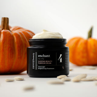 A black jar of Savor Beauty Enchant Sleeping Beauty Pumpkin Cream with white cream visible on top, styled on a white surface with pumpkin seeds in the foreground and orange pumpkins blurred in the background.