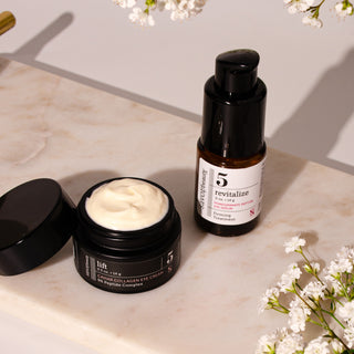 an open black jar of Caviar Collagen Eye Cream showing the white cream inside, and a black pump bottle of Revitalize Eye Cream, styled on a marble surface with white flowers.