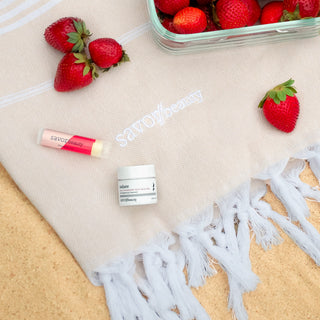 Cosmetic products and strawberries on a beige towel with 'Savor Beauty' branding.