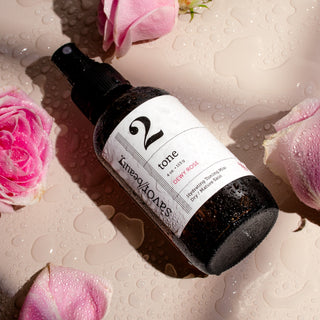 Wet dark spray bottle with white label reading '2' laying on a neutral background surrounded by pink roses.