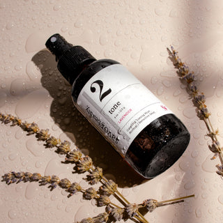 Wet dark spray bottle with white label reading '2' laying on a neutral background surrounded by sprigs of lavender.