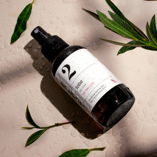 Wet dark spray bottle with white label reading '2' laying on a neutral background surrounded by leaves.