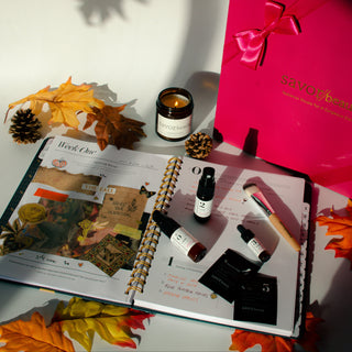 Open planner with beauty products, pink box, and autumn leaves on a white surface.
