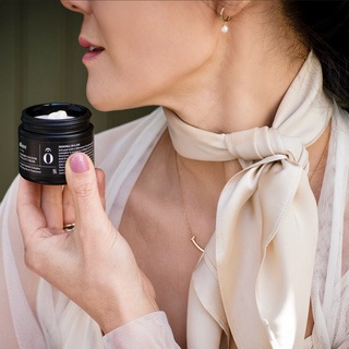 Woman holding a black jar with a white label, wearing a beige scarf and gold necklace.