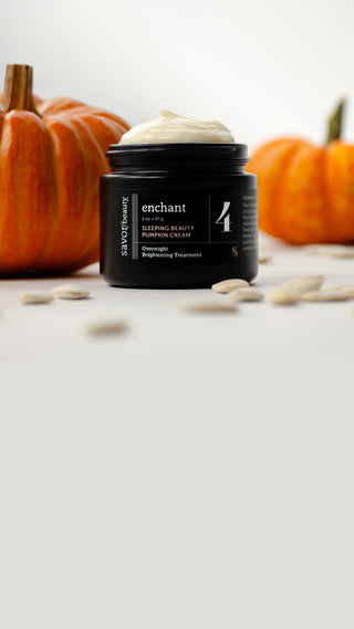 A black jar of Enchant Sleeping Beauty Pumpkin Cream with white cream on top, styled with pumpkins and pumpkin seeds on a light gray surface.