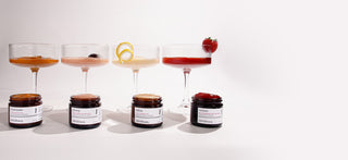 Four savor beauty peels lined up on a white surface, each paired with a stemmed coupe-style glass containing a different colored peel textures: orange, peach, pale yellow, and red. 