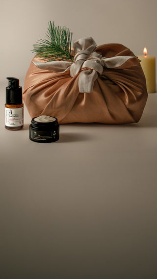 A styled product shot featuring a brown fabric gift bag tied with a beige ribbon and decorated with pine needles, alongside a small amber bottle of pomegranate eye serum and a black jar of caviar collagen eye cream, set against a warm beige background with a lit candle.
