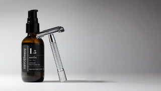 Dark amber and black label bottle of Savor Beauty's Sapphire Post-Shave Gel on a gray clean background with an aluminum straight razor leaning on the bottle