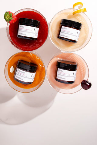 Four savor beauty peels lined up on a white surface, each paired with a stemmed coupe-style glass containing a different colored peel textures: orange, peach, pale yellow, and red. 