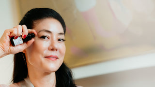A woman with dark hair applies a cosmetic product from a small black tube held by a hand with red nail polish. The background is a softly blurred warm-toned interior.